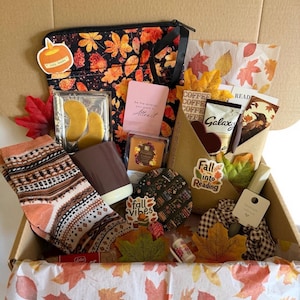May include: A gift box containing a black pouch with orange autumn leaves, a pair of patterned socks, a brown mug, gold eye masks, a Galaxy chocolate bar, a bookmark, a scrunchie, and various stickers with text including Fall into Reading.