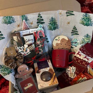 Puede incluir: Una caja de regalo con temática navideña llena de artículos festivos. Incluye una taza roja, calcetines con renos, un hombre de jengibre, chocolate instantáneo Galaxy, una scrunchie y otras golosinas navideñas. La caja está forrada con papel con árboles de Navidad.
