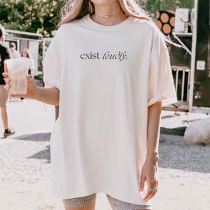 May include: Cream-colored oversized t-shirt with the phrase "exist loudly" printed in a dark, elegant font. The shirt has short sleeves and a relaxed fit. The model is wearing the shirt outdoors.