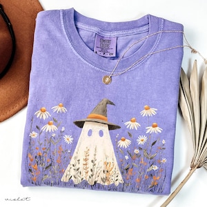 May include: A violet Comfort Colors t-shirt featuring a whimsical ghost wearing a witch hat, surrounded by daisies and wildflowers. A gold necklace with a pendant rests on the shirt. The shirt is folded, with a brown hat and a dried palm leaf in the background.
