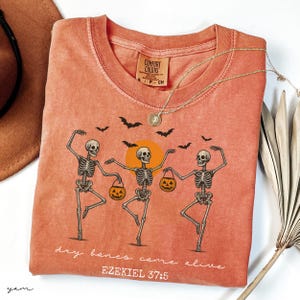 May include: Coral-colored t-shirt featuring a graphic of three dancing skeletons holding jack-o'-lanterns, with bats and a sun graphic. The text "dry bones come alive" and "EZEKIEL 37:5" are printed on the shirt. A gold necklace is draped across the shirt.