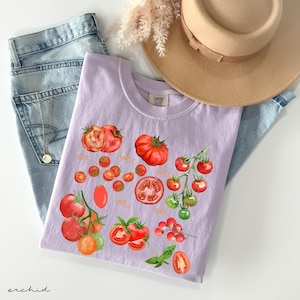 May include: A light purple t-shirt with a watercolor design of red tomatoes. The shirt has the text "COMFORT COLORS" on the neck.
