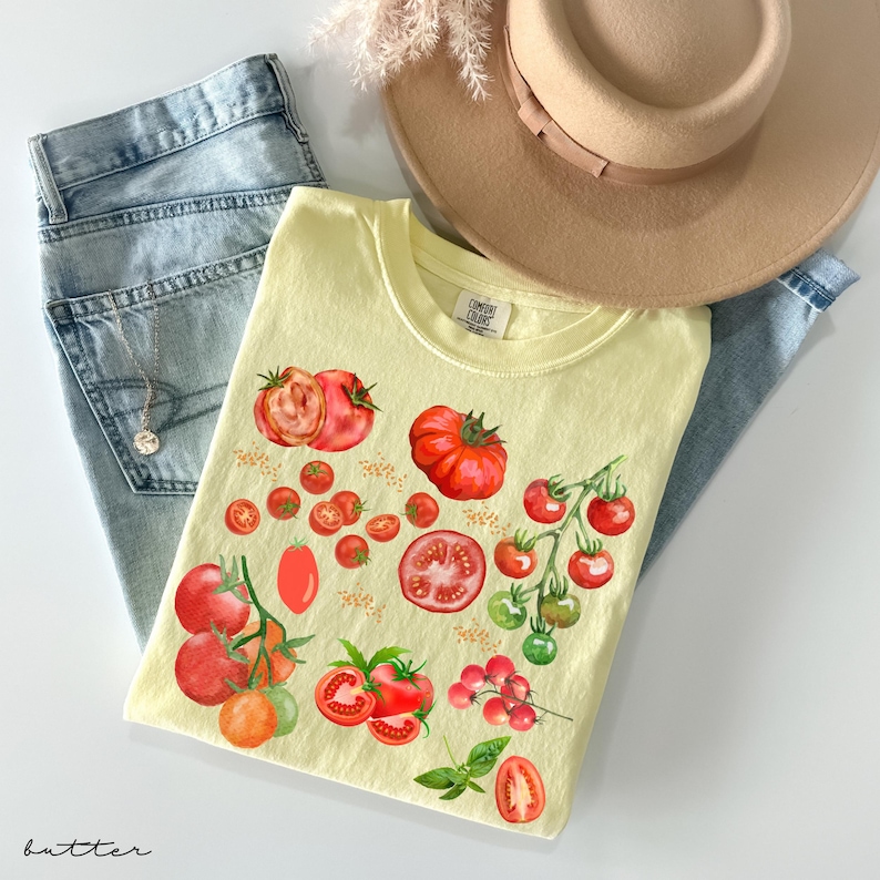 May include: A yellow t-shirt with a watercolor design of red tomatoes. The shirt has a small tag that says "Canvas Craft" on it. The shirt is laid out on a blue denim background.