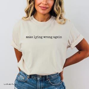 May include: Ivory-colored t-shirt with the phrase "make lying wrong again" printed in black. The shirt has short sleeves and is paired with blue denim shorts. The model is wearing the shirt.