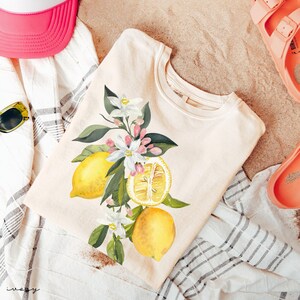 May include: A cream-coloured t-shirt with a watercolour illustration of lemons, blossoms, and leaves. The design features whole and sliced lemons with white flowers and green foliage. The shirt is on a striped towel with a pink hat and sandals.