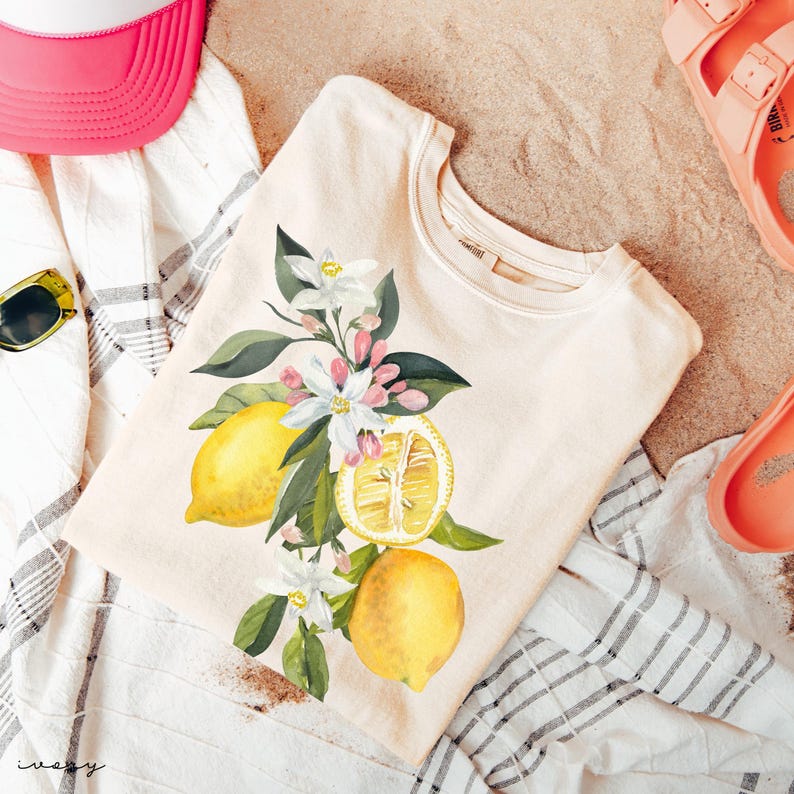 May include: A cream-coloured t-shirt with a watercolour illustration of lemons, blossoms, and leaves. The design features whole and sliced lemons with white flowers and green foliage. The shirt is on a striped towel with a pink hat and sandals.