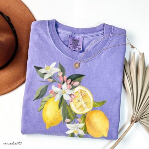 May include: A violet Comfort Colors t-shirt with a lemon and floral design. The graphic features yellow lemons, white flowers, and green leaves. A gold necklace with a pendant rests on the shirt.