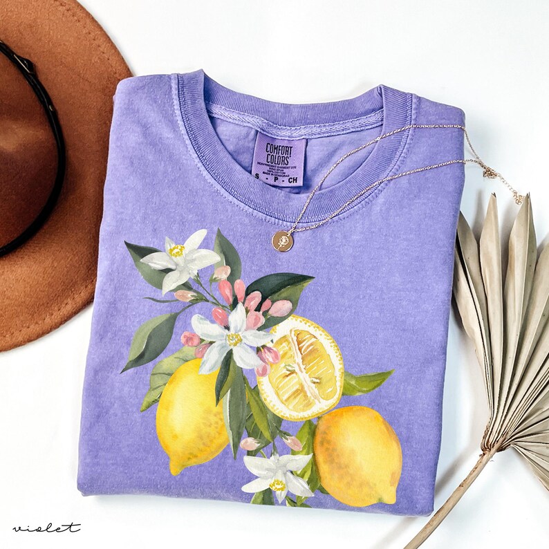 May include: A violet Comfort Colors t-shirt with a lemon and floral design. The graphic features yellow lemons, white flowers, and green leaves. A gold necklace with a pendant rests on the shirt.