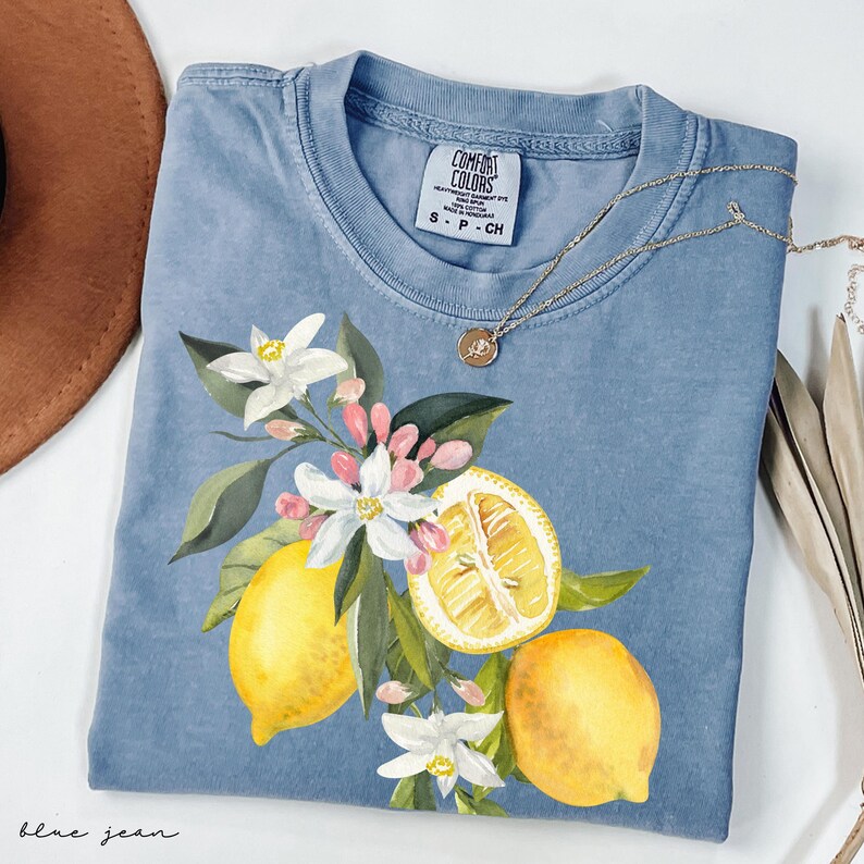 May include: A blue jean Comfort Colors t-shirt with a watercolour design of lemons, white flowers, and pink buds. A gold necklace with a pendant rests on the shirt. The shirt is labelled S-P-CH.