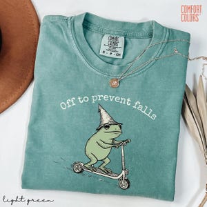 May include: Light green t-shirt with the text "Off to prevent falls" above a cartoon frog riding a scooter and wearing a wizard hat. The shirt has a crew neck and short sleeves. A silver necklace with a pendant is draped across the shirt.