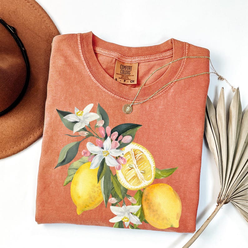 May include: Coral Comfort Colors t-shirt with a watercolour design of lemons, white flowers, and green leaves. A gold necklace with a pendant rests on the shirt. A brown hat and dried palm leaf are in the background.