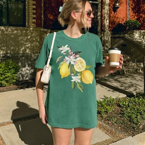 May include: A green t-shirt with a watercolour design of lemons, white blossoms, and green leaves. The design features whole and sliced lemons. The shirt is a short-sleeved, casual style.