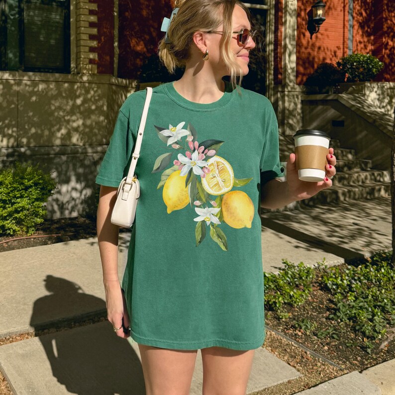 May include: A green t-shirt with a watercolour design of lemons, white blossoms, and green leaves. The design features whole and sliced lemons. The shirt is a short-sleeved, casual style.