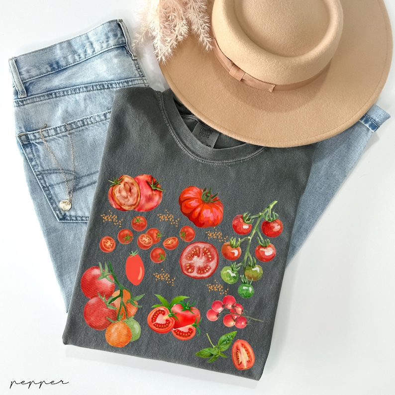 May include: A dark gray t-shirt with a graphic design of red tomatoes, green leaves, and tomato seeds. The shirt is laid out on a pair of blue jeans and a brown hat.