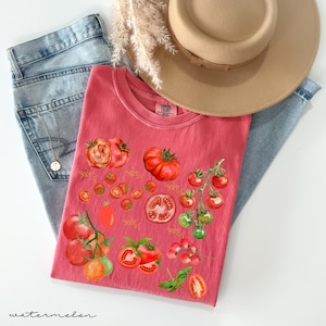May include: A red t-shirt with a watercolor design of tomatoes, including whole tomatoes, tomato slices, and cherry tomatoes. The shirt has a distressed look and a tag that says "Comfy Colors".