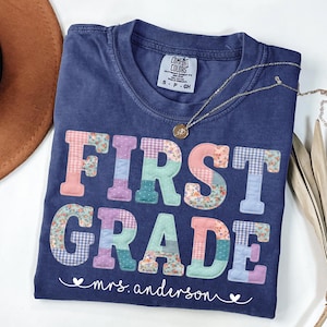 May include: A navy blue t-shirt featuring the text FIRST GRADE in large, multi-colored patchwork-style letters with floral and gingham patterns. Below the text, the name mrs. anderson is printed in white script with small heart accents.