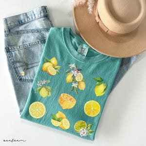 May include: A teal t-shirt with a lemon design, paired with light-wash jeans, a gold necklace, and a tan hat. The shirt features watercolor illustrations of lemons, lemon slices, and blossoms. The word "seafarm" is written in cursive.