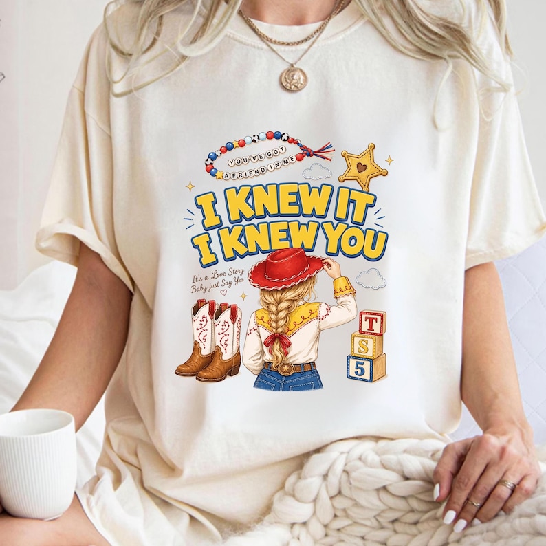 K&ouml;nnte beinhalten: Ein cremefarbenes T-Shirt mit einem Grafikdruck einer Person mit blondem Zopf, die einen roten Cowboyhut, braune Stiefel und ein gelbes Hemd tr&auml;gt. Der Text lautet I KNEW IT I KNEW YOU, It's a Love Story Baby just Say Yes und YOU'VE GOT A FRIEND IN ME.