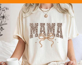 May include: Cream-colored t-shirt with the word "MAMA" in a leopard print design. A tan bow and small stars are below the word. The text "Custom DTF Transfer" is in a purple font at the top.
