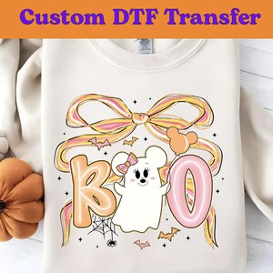 May include: Cream-colored sweatshirt with a Halloween-themed graphic. The design features the word "BOO" in orange and pink bubble letters, a cartoon ghost, bats, a spiderweb, and a large bow. The text "Custom DTF Transfer" is at the top.