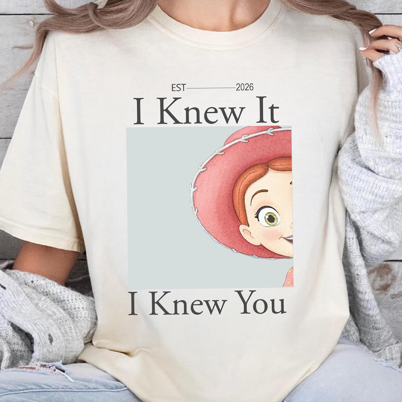 May include: A cream-colored short-sleeve t-shirt featuring the text "EST 2026 I Knew It I Knew You" in dark gray lettering. A graphic of a cartoon character with red hair and a pink cowboy hat is centered on the shirt, partially visible on the right side.