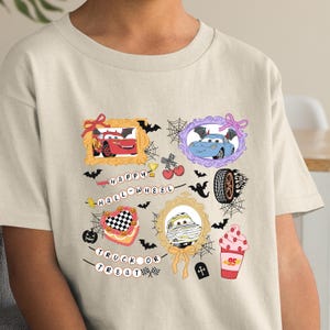 May include: A light beige t-shirt featuring a colorful Halloween-themed graphic. The design includes framed images of cartoon cars, bats, spiderwebs, and the text "HAPPY HALL-WHEEL" and "TRUCK OR TREAT".