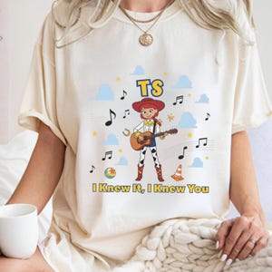 K&ouml;nnte beinhalten: Ein cremefarbenes T-Shirt mit einer Grafik einer Cowgirl-Figur, die eine akustische Gitarre spielt. Das Design enth&auml;lt blaue Wolken, gelbe Sterne, Musiknoten, einen Ball, einen Verkehrskegel sowie den Text TS und I Knew It, I Knew You.