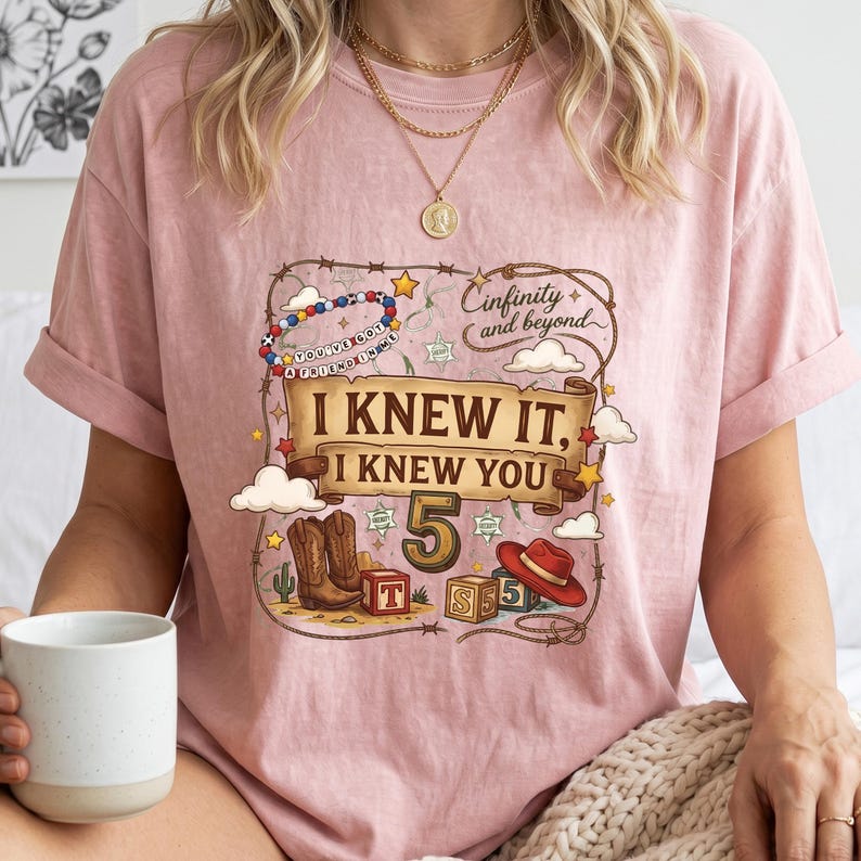 K&ouml;nnte beinhalten: Ein altrosa T-Shirt mit einem Grafikdruck, der von einem Lasso umrandet ist, einem Banner mit der Aufschrift "I KNEW IT, I KNEW YOU", der Zahl "5", Cowboystiefeln, einem roten Hut, Alphabet-W&uuml;rfeln sowie dem Text "Cinfinity and beyond" und "YOU'VE GOT A FRIEND IN ME".