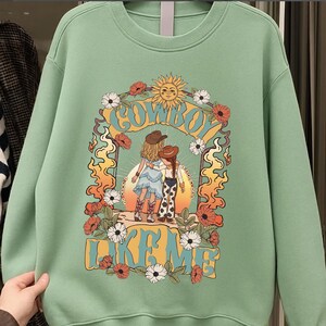 May include: A light green crewneck sweatshirt featuring a graphic print of two figures in cowboy hats and boots standing under a sun archway. The text "COWBOY LIKE ME" is printed in large yellow letters, surrounded by orange flames and white and red flowers.
