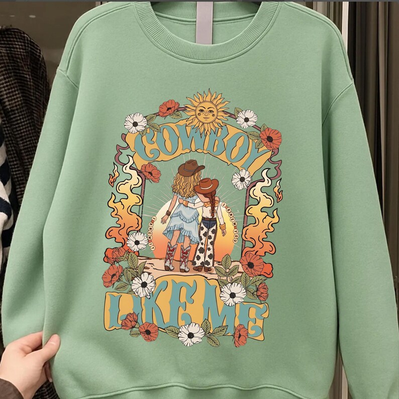 May include: A light green crewneck sweatshirt featuring a graphic print of two figures in cowboy hats and boots standing under a sun archway. The text "COWBOY LIKE ME" is printed in large yellow letters, surrounded by orange flames and white and red flowers.