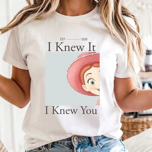 May include: A white short-sleeved t-shirt featuring a graphic of a cartoon character wearing a pink hat, partially visible within a light blue square. The text "EST 2026", "I Knew It", and "I Knew You" is printed in dark gray lettering above and below.