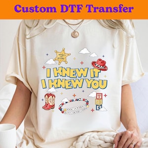 Puede incluir: Una camiseta color crema con un estampado gr&aacute;fico que incluye el texto I KNEW IT I KNEW YOU. El dise&ntilde;o presenta una estrella de sheriff amarilla, un sombrero de vaquero rojo, botas de vaquero rojas, una pulsera de cuentas y bloques.