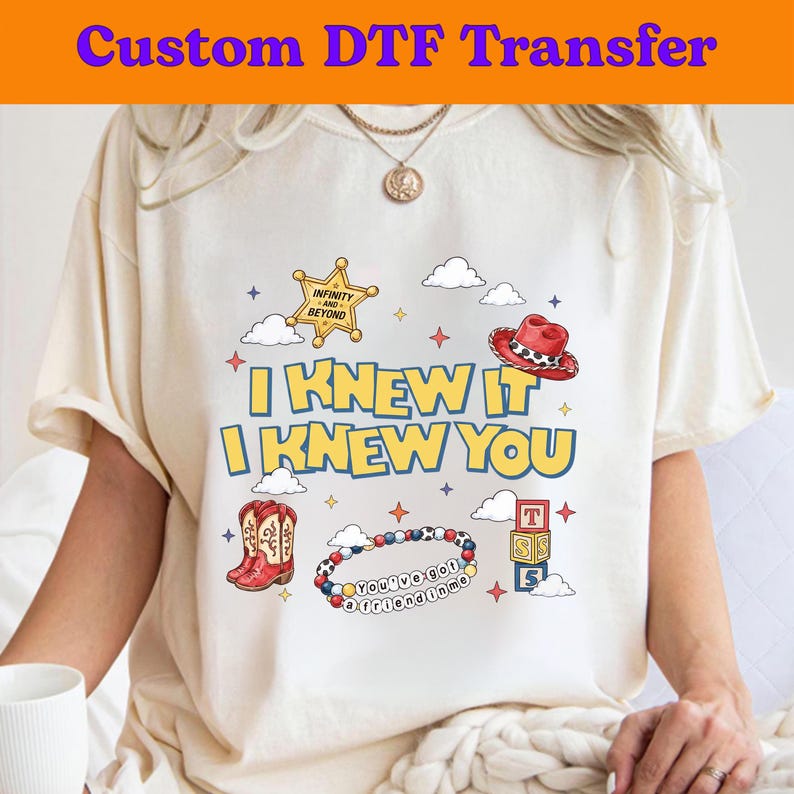 Puede incluir: Una camiseta color crema con un estampado gr&aacute;fico que incluye el texto I KNEW IT I KNEW YOU. El dise&ntilde;o presenta una estrella de sheriff amarilla, un sombrero de vaquero rojo, botas de vaquero rojas, una pulsera de cuentas y bloques.