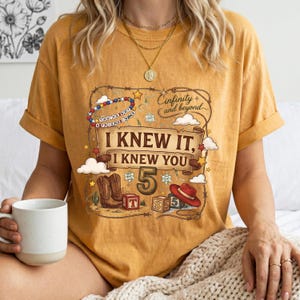K&ouml;nnte beinhalten: Ein senfgelbes T-Shirt mit einem Grafikdruck, der ein Schriftband mit dem Text I KNEW IT, I KNEW YOU, die Zahl 5, einen Cowboyhut, Stiefel, Spielw&uuml;rfel und den Schriftzug Infinity and beyond sowie ein Lasso und kleine Sterne zeigt.