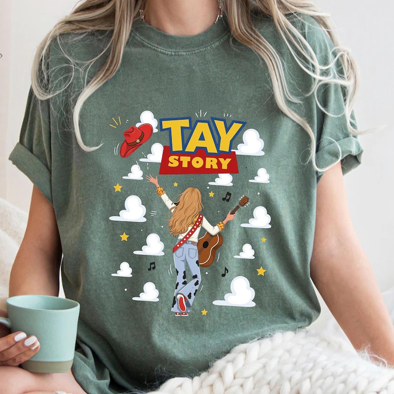 K&ouml;nnte beinhalten: Ein salbeigr&uuml;nes T-Shirt mit einer Grafik einer Person mit langem Haar, die eine Akustikgitarre spielt, eine Hose mit Kuhmuster und einen roten Cowboyhut tr&auml;gt. Der Text TAY STORY ist in fetten gelben und roten Buchstaben aufgedruckt.