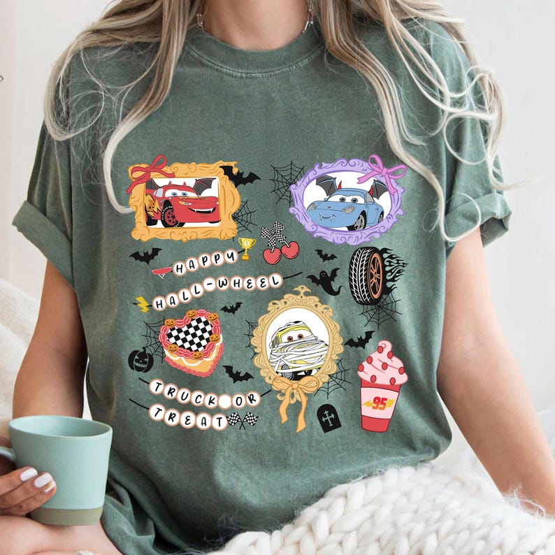 May include: Green t-shirt featuring Halloween-themed graphics of cartoon cars in frames, bats, spiderwebs, and the phrases "Happy Hall-Wheel" and "Truck or Treat". The design includes a checkered heart and a mummy car. A cup is visible in the lower left corner.