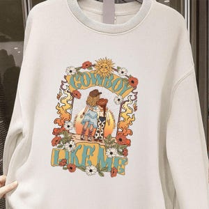 May include: A cream-colored crewneck sweatshirt featuring a graphic print of two figures in cowboy hats and boots standing before a sun. The design includes floral accents, flame-like borders, and the text "COWBOY LIKE ME" in bold blue lettering.