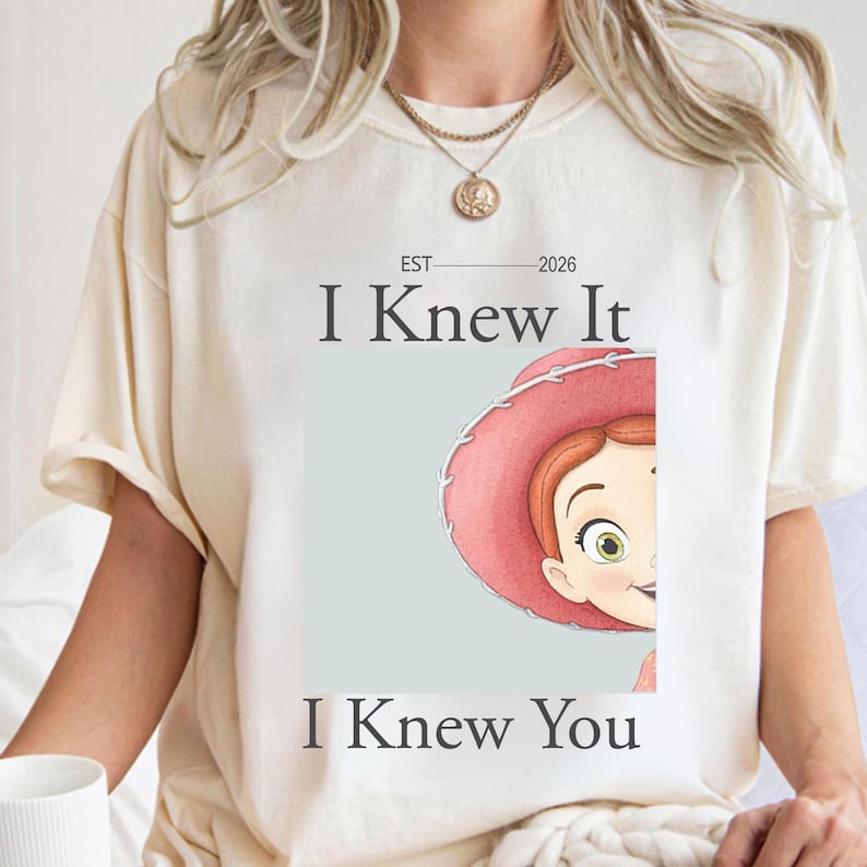 May include: A cream-colored short-sleeve t-shirt featuring a graphic of a cartoon character wearing a pink cowboy hat. The text reads EST 2026, I Knew It, and I Knew You. The design is centered on the chest of the shirt, which has a crew neckline.