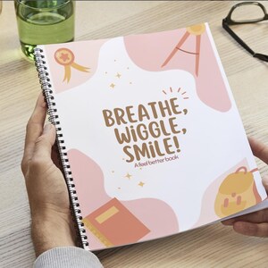 Op de afbeelding: Een spiraalgebonden notitieboek met de tekst "Breathe, Wiggle, Smile!" en "A feel better book" op de kaft. Het kaftontwerp bevat roze vormen, sterren en illustraties van een medaille, kompas, boek en rugzak.