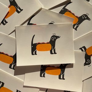 May include: A collection of white cards featuring a black dachshund dog illustration with an orange hot dog bun body and a red zigzag detail. The dog has a small white highlight on its head and nose.