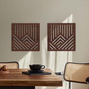 May include: Two square, wooden wall art pieces with geometric designs. Each piece features a dark brown frame with vertical and angled cutouts, creating a layered effect. The art is mounted on a neutral-colored wall, adding a modern touch to the space.