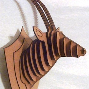 Puede incluir: Escultura de pared de madera con forma de cabeza de órix. La escultura está hecha de madera en capas, con rayas marrones oscuras en la cabeza y los cuernos. Los cuernos se curvan hacia arriba. Montada sobre una placa de madera en forma de escudo.