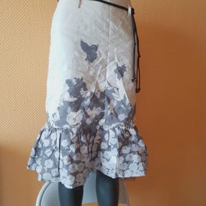 Könnte beinhalten: Ein cremefarbener Rock mit floralem Muster und Rüschensaum. Der Rock hat einen schwarzen Gürtel und ein dekoratives Blumendesign in Grau und Weiß. Das Wort "Atelier" ist auf dem Bund aufgedruckt.