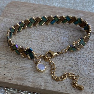 Peut inclure: Un bracelet doré avec un motif tissé de petites perles rectangulaires vertes, bronze et bleues. Un petit charme rond rose pâle est suspendu à la fermeture. Le bracelet est présenté sur une surface en bois.