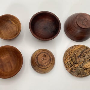 May include: Six handcrafted wooden bowls in various shades of brown. The bowls are round with varying depths and wood grain patterns. Some bowls have markings on the bottom. A natural, rustic aesthetic.