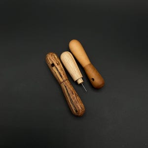 May include: Three wooden tools with rounded handles. The tools are in various shades of brown, with one featuring a striped pattern. One tool has a needle-like tip, while another has a hole. The tools are arranged on a dark background.
