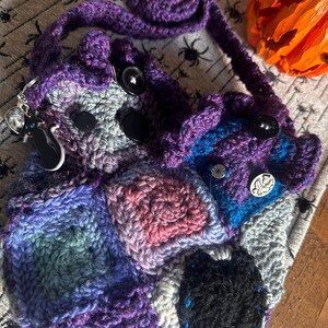 May include: A handmade crochet bag in shades of purple, blue, gray, and pink. The bag features a patchwork design with black buttons and a long strap. A black cat keychain hangs from the side. A decorative pumpkin is in the background.