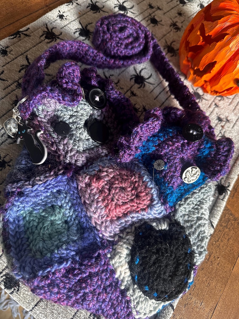 May include: A handmade crochet bag in shades of purple, blue, gray, and pink. The bag features a patchwork design with black buttons and a long strap. A black cat keychain hangs from the side. A decorative pumpkin is in the background.