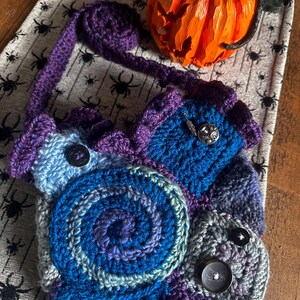 May include: A handmade crochet bag in shades of blue, purple, and gray, featuring a spiral design and button closures. The bag has a long purple strap and is likely a shoulder bag or crossbody bag. The bag is on a table with a Halloween theme.