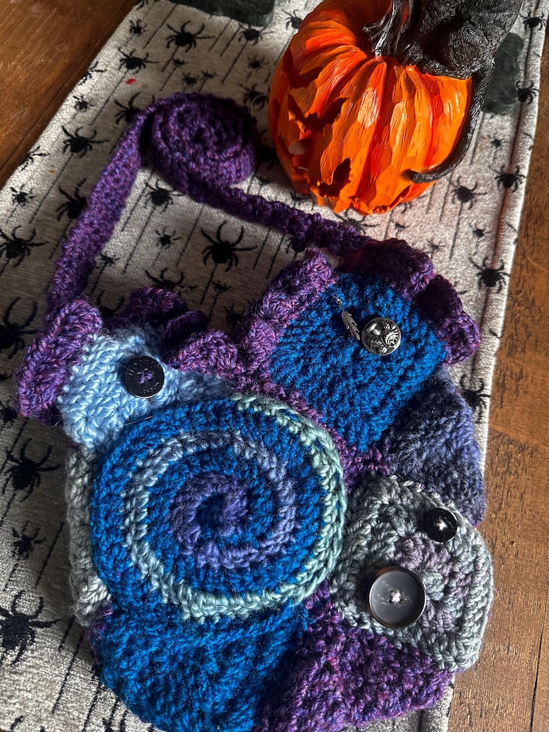 May include: A handmade crochet bag in shades of blue, purple, and gray, featuring a spiral design and button closures. The bag has a long purple strap and is likely a shoulder bag or crossbody bag. The bag is on a table with a Halloween theme.